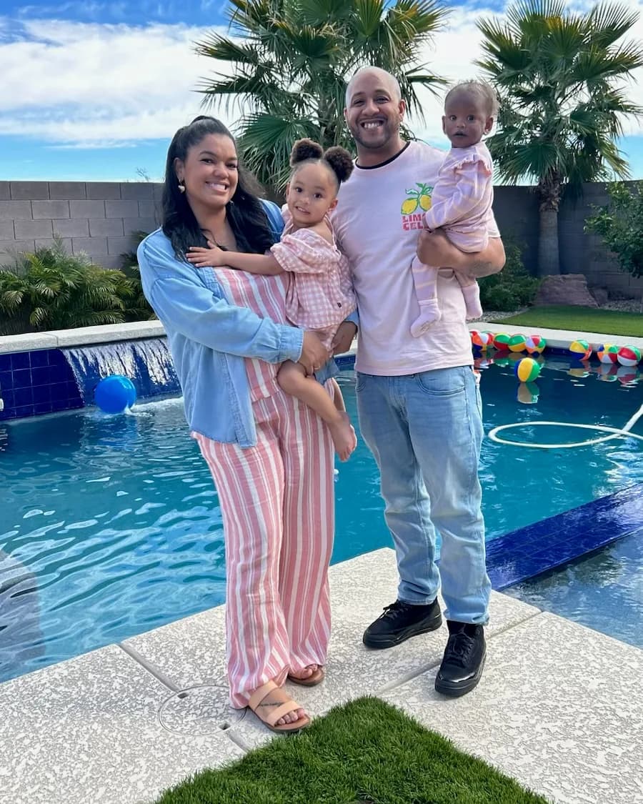 Josh and Shade’ smiling by a backyard pool under palm trees, each holding one of their two young children