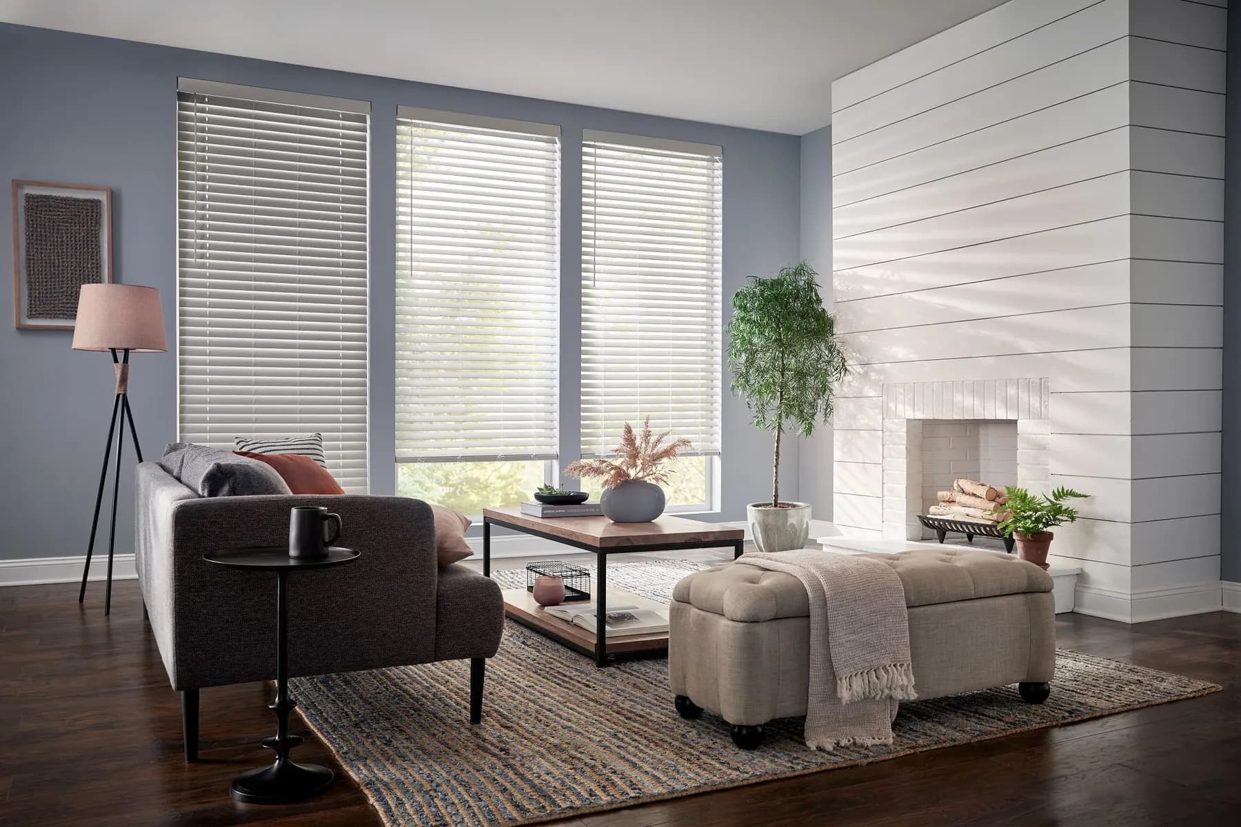 White faux wood blinds on three windows of a blue-grey living room with a white shiplap fireplace.