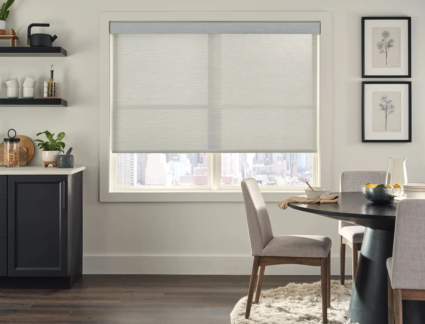 A light grey roller shade half lowered on a breakfast-nook window with a round dining table and a city view.