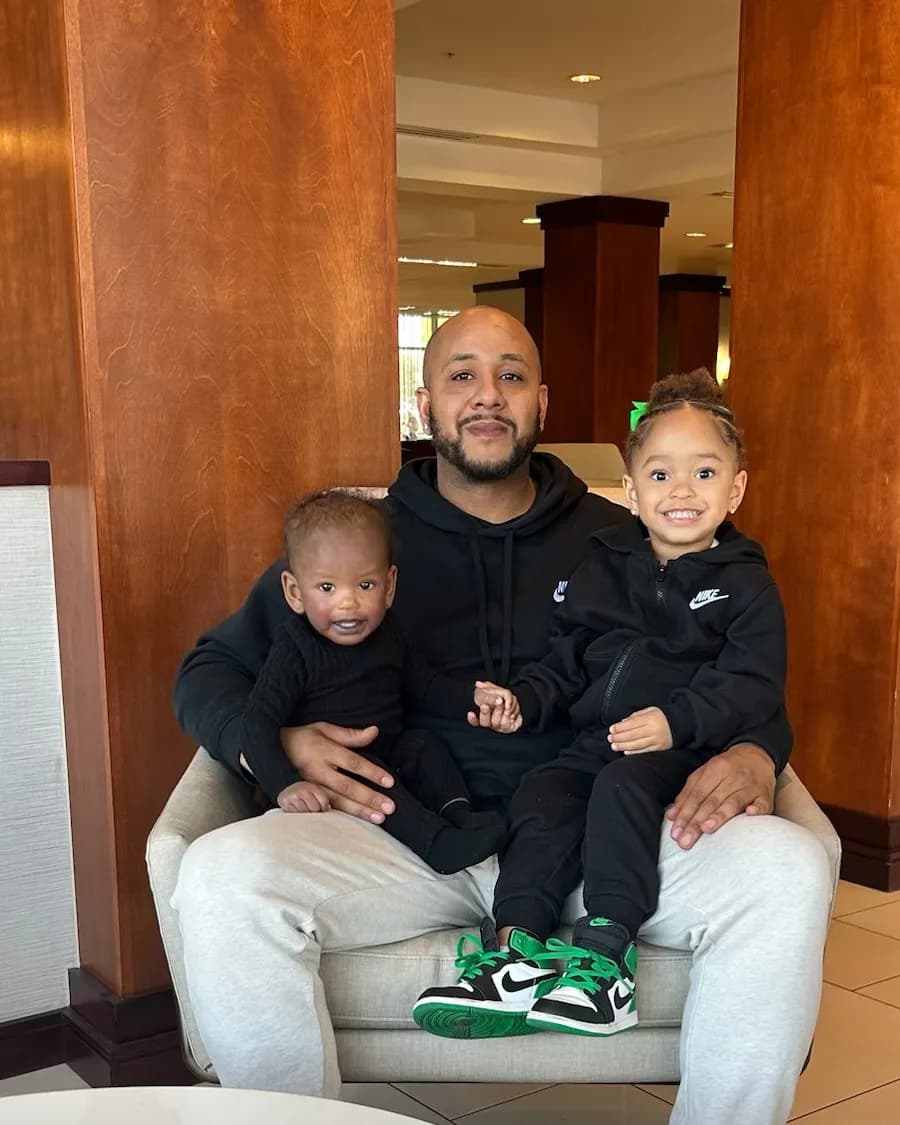 Josh sitting in an armchair with their baby son and young daughter on his lap, all three in black, the daughter smiling in green sneakers