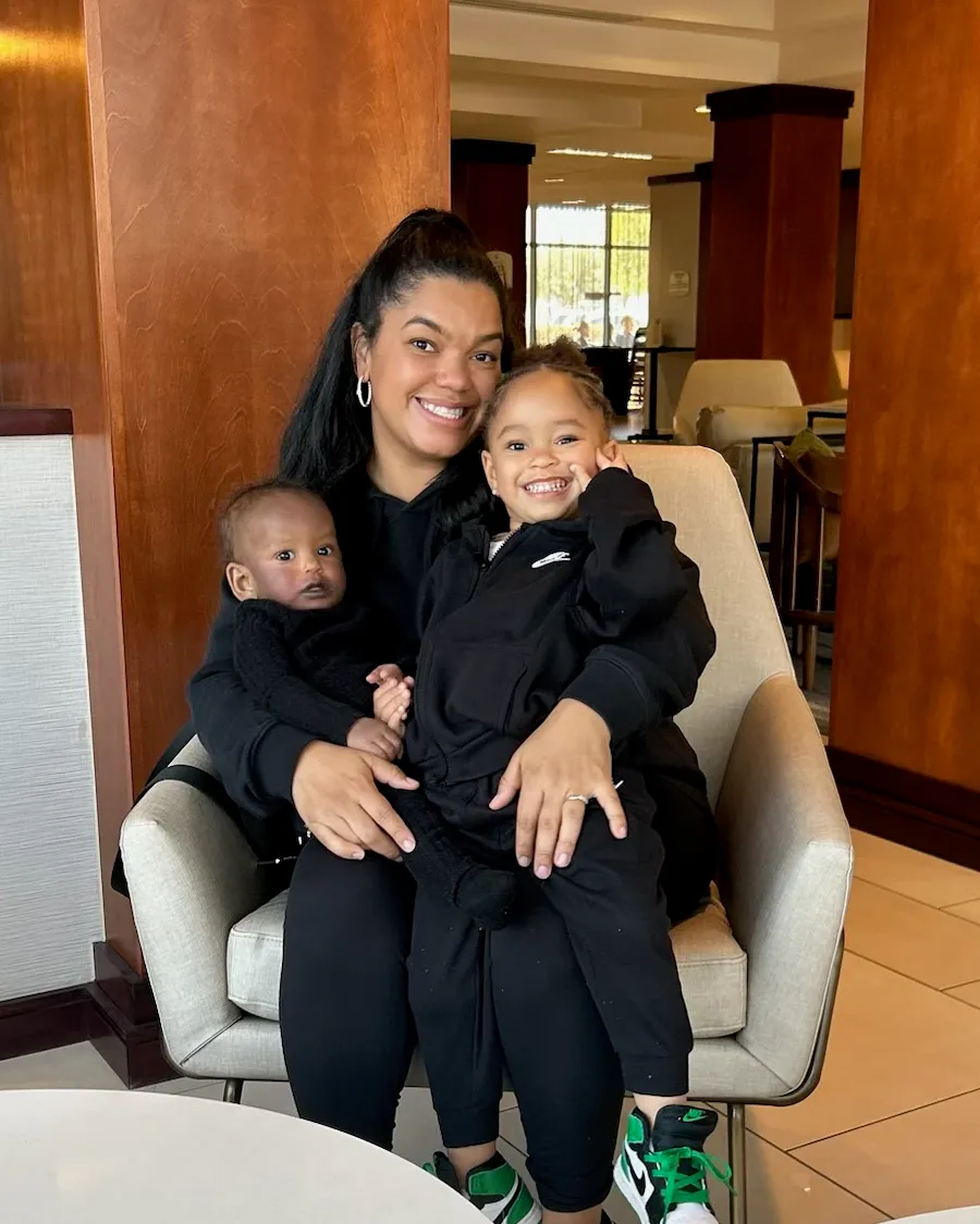 Shade’ smiling in an armchair with their baby son and young daughter on her lap, the daughter grinning with a hand to her cheek