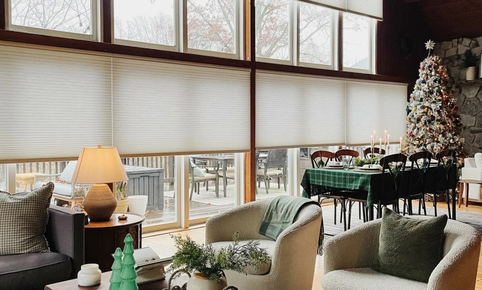 White cellular shades lowered partway across a wall of tall windows in a wood-paneled great room, with armchairs and a holiday-set dining table in front.