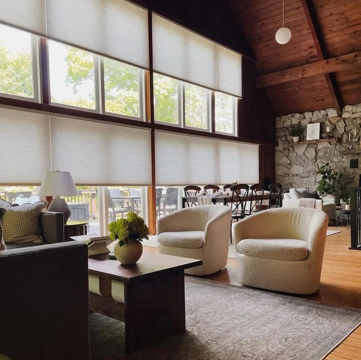 White cellular shades lowered across two rows of windows in a vaulted great room with exposed wood beams and a stone fireplace.