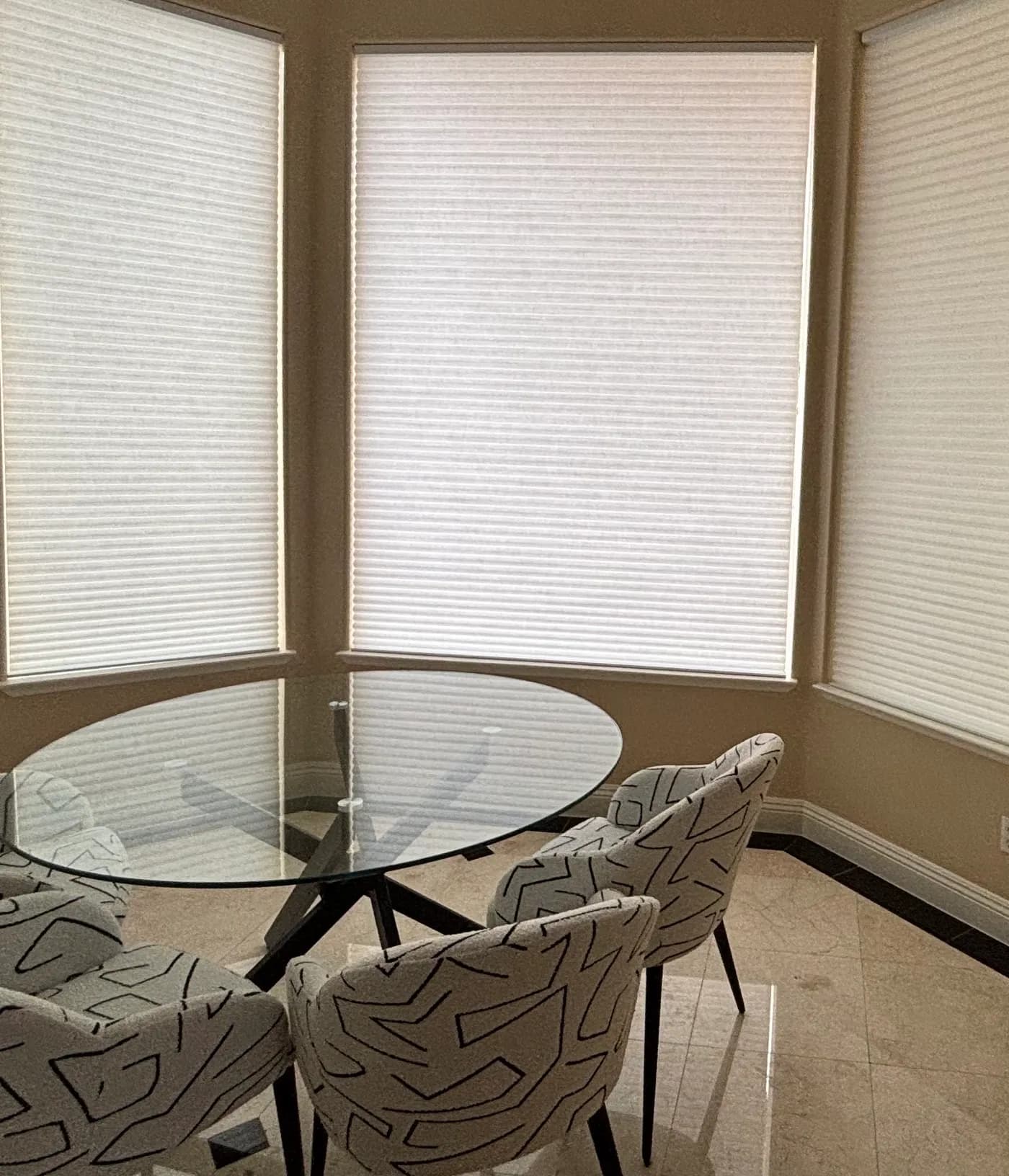 Motorized white cellular shades lowered on the windows of a bay breakfast nook, around a round glass table with patterned upholstered chairs.