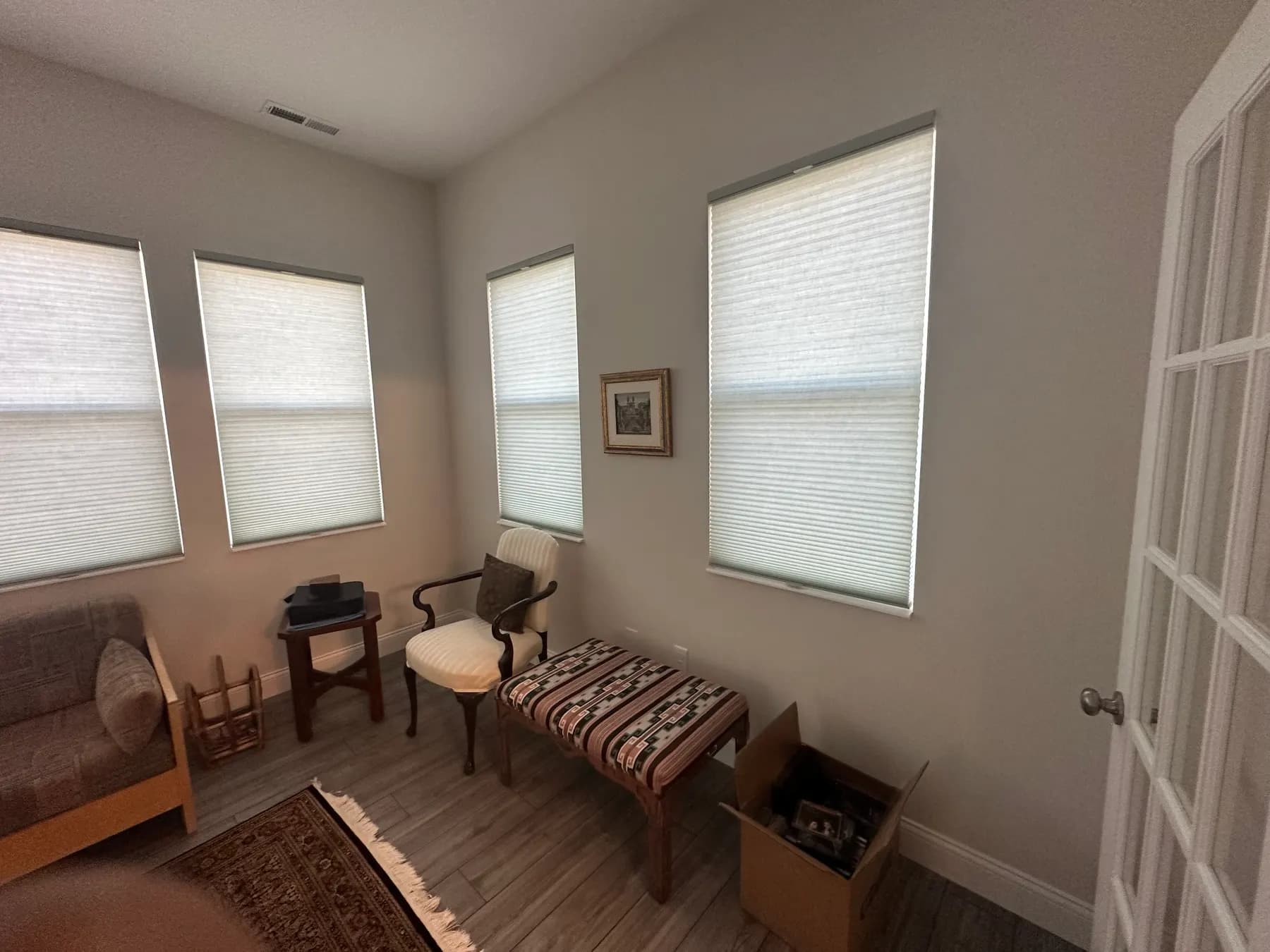 Light cellular shades lowered on four windows in a sitting room, filtering afternoon sun onto a wood floor.