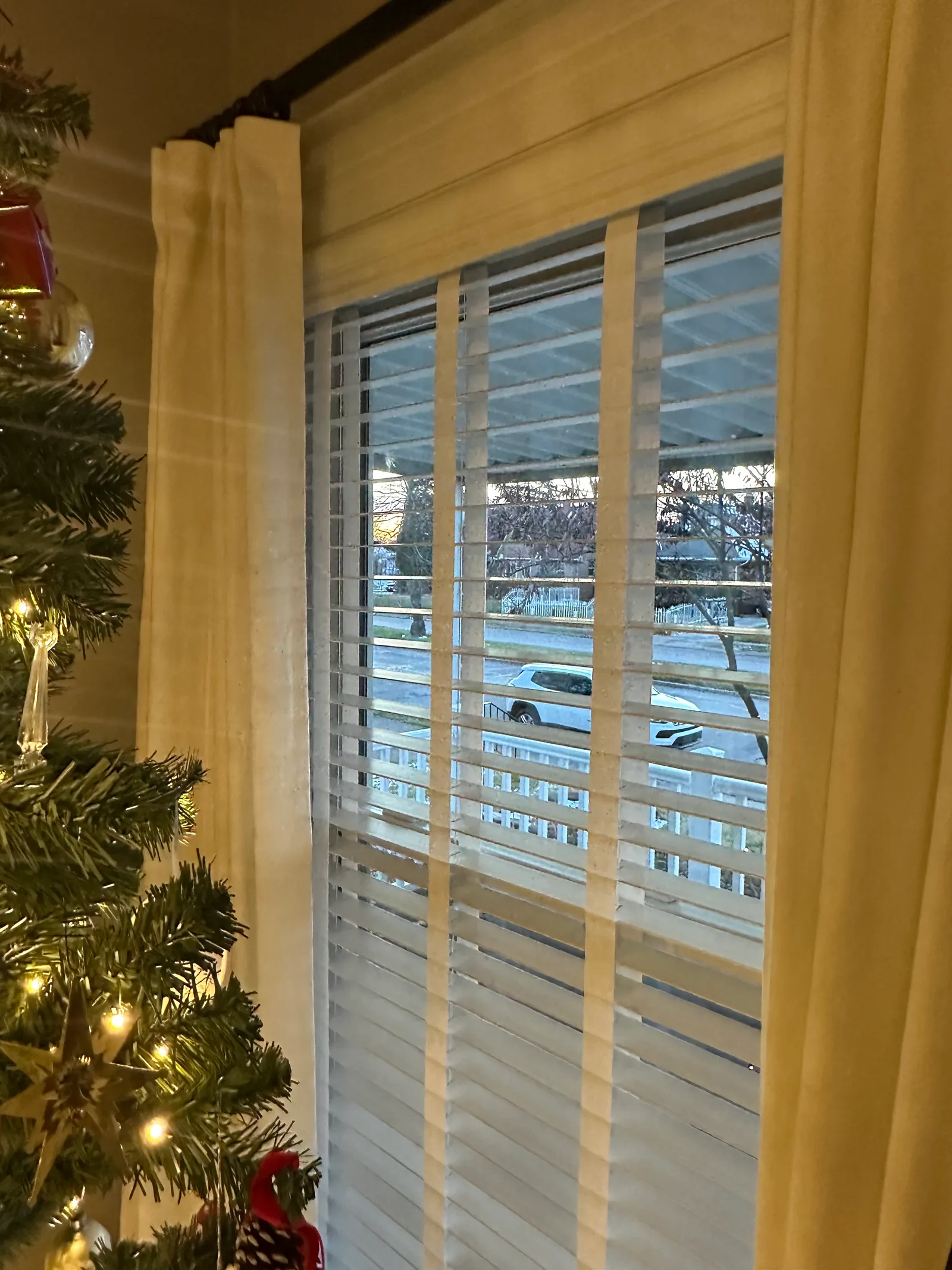 White faux wood blinds with wide slats and cloth tapes in a tall front window, framed by cream curtains, looking out to a covered porch and the street.