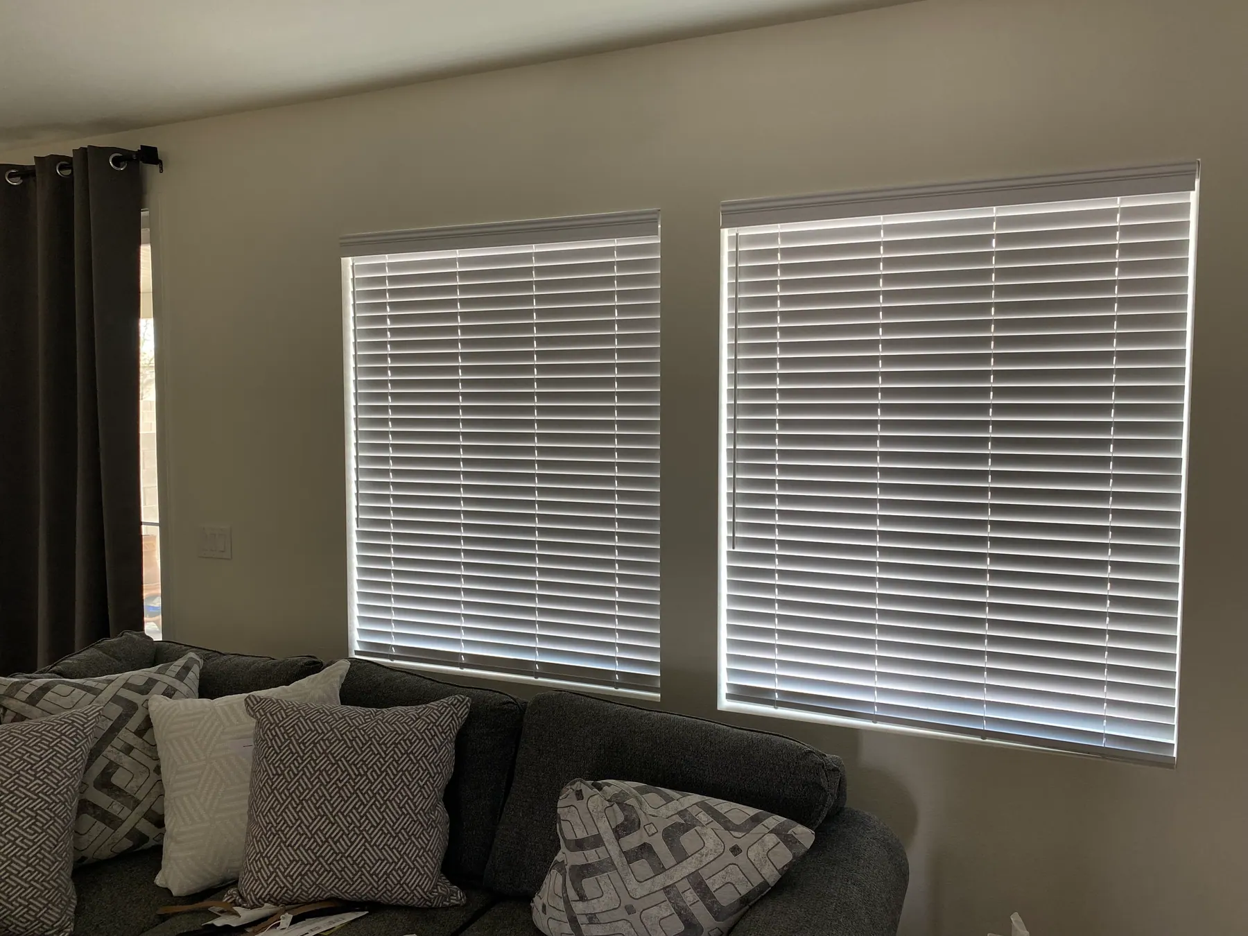 White faux wood blinds with wide slats on two windows above a grey sofa with patterned pillows.