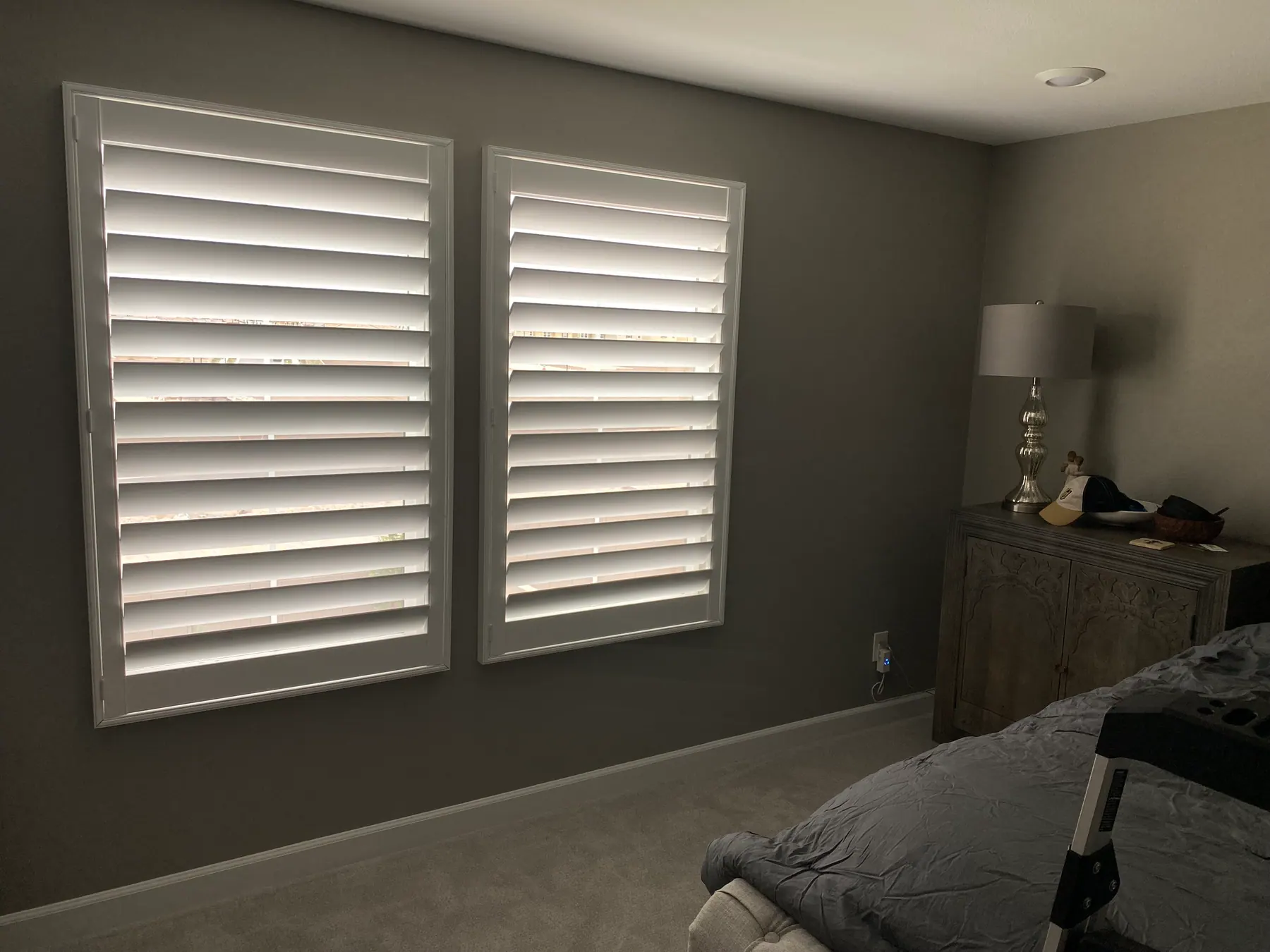 White plantation shutters with open louvers on two windows in a bedroom with grey walls.