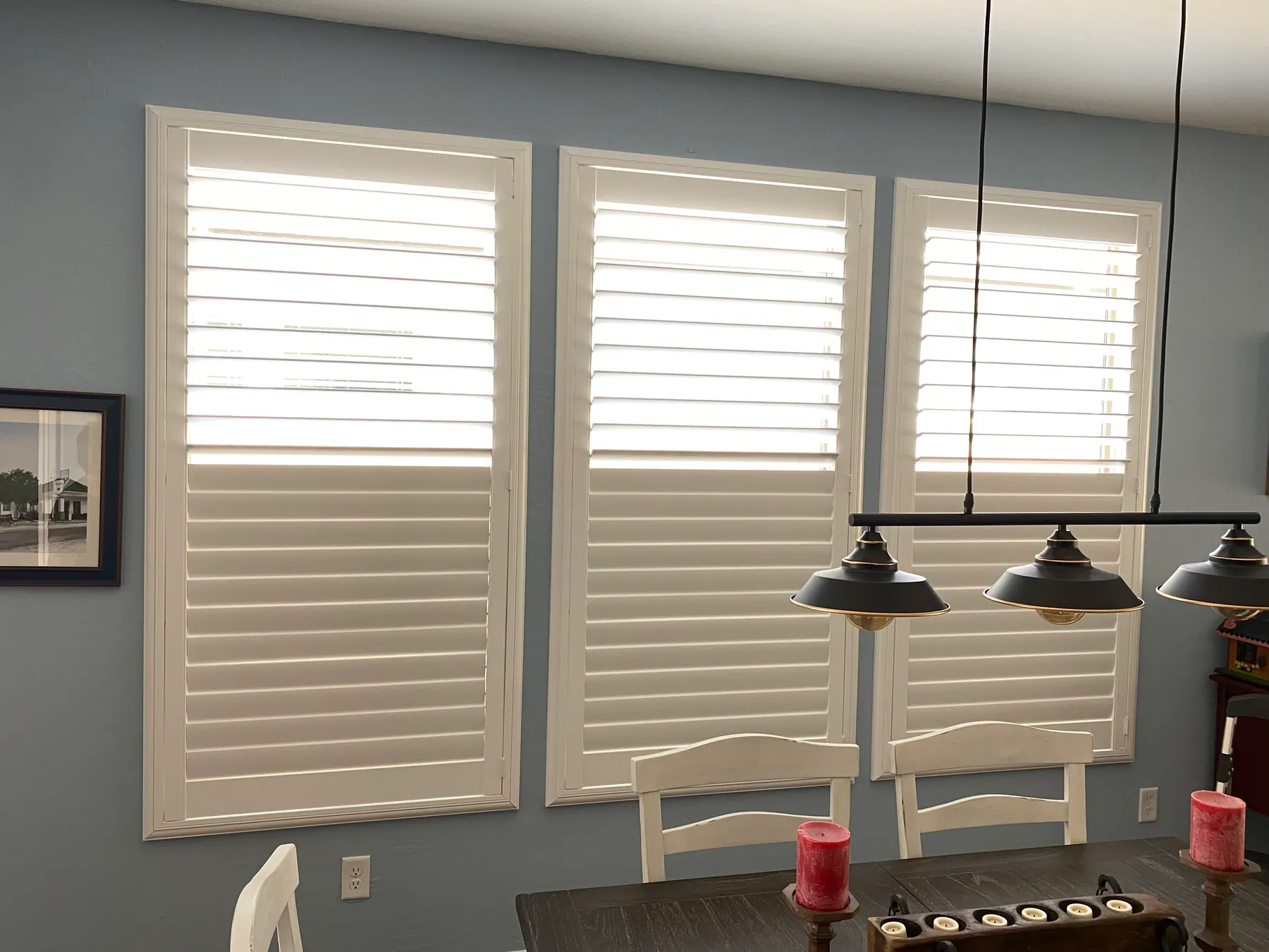 White plantation shutters on three windows in a dining room with blue walls and industrial pendant lights over the table.