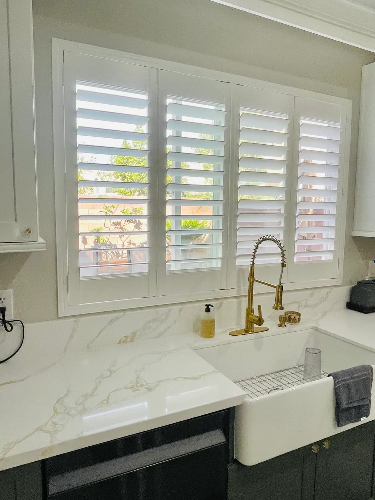 White plantation shutters with open louvers over a farmhouse kitchen sink and marble countertop