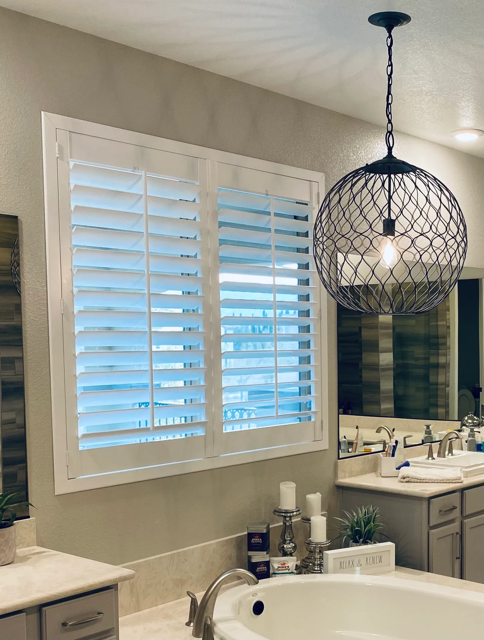 White plantation shutters with louvers tilted open on a wide primary-bathroom window, beside a round wire pendant light above the tub.