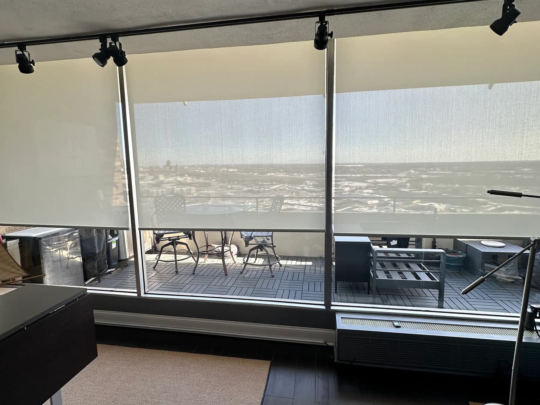 A sheer roller shade lowered across a wall of glass in a bedroom, with a deck and a lake visible through the fabric.