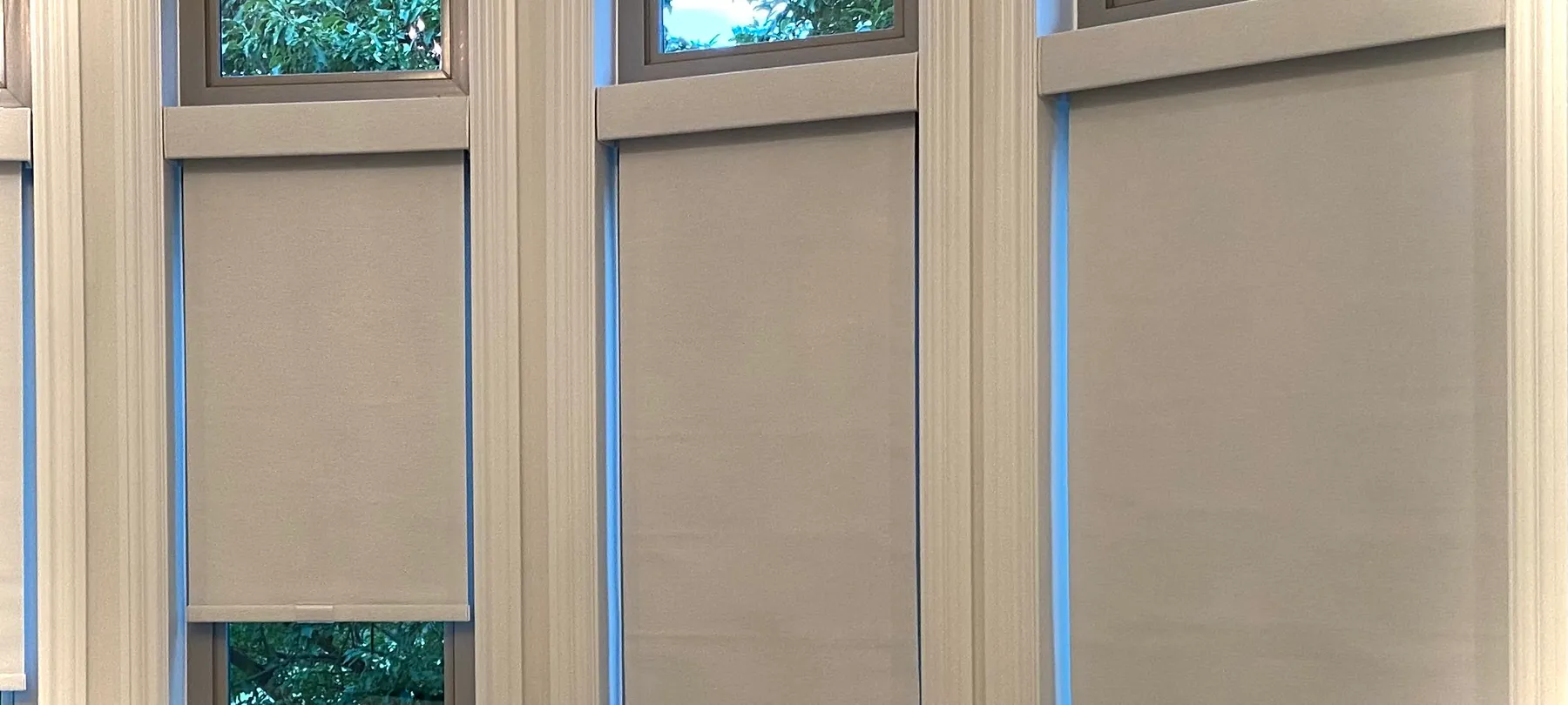 Close view of light grey roller shades in a white-trimmed bay window, one raised to show trees below, with clear transom windows above.