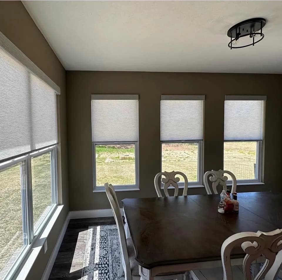 Light textured roller shades in slim cassettes, half raised on four windows around a dining table, with a green yard outside.
