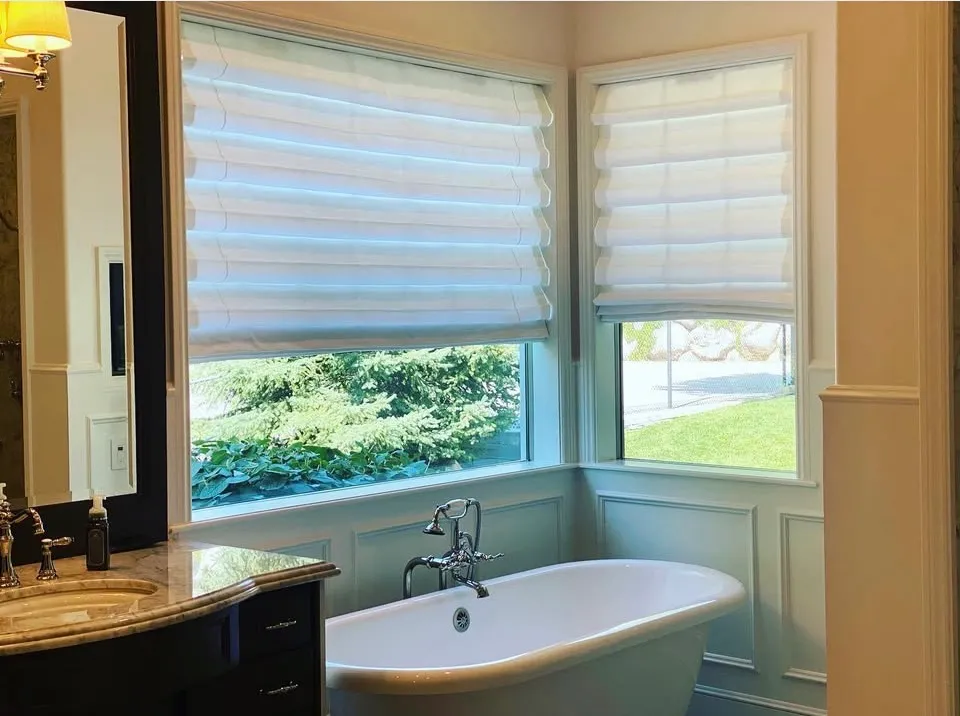 White hobbled roman shades in soft folds on two corner windows above a freestanding tub in a primary bathroom.
