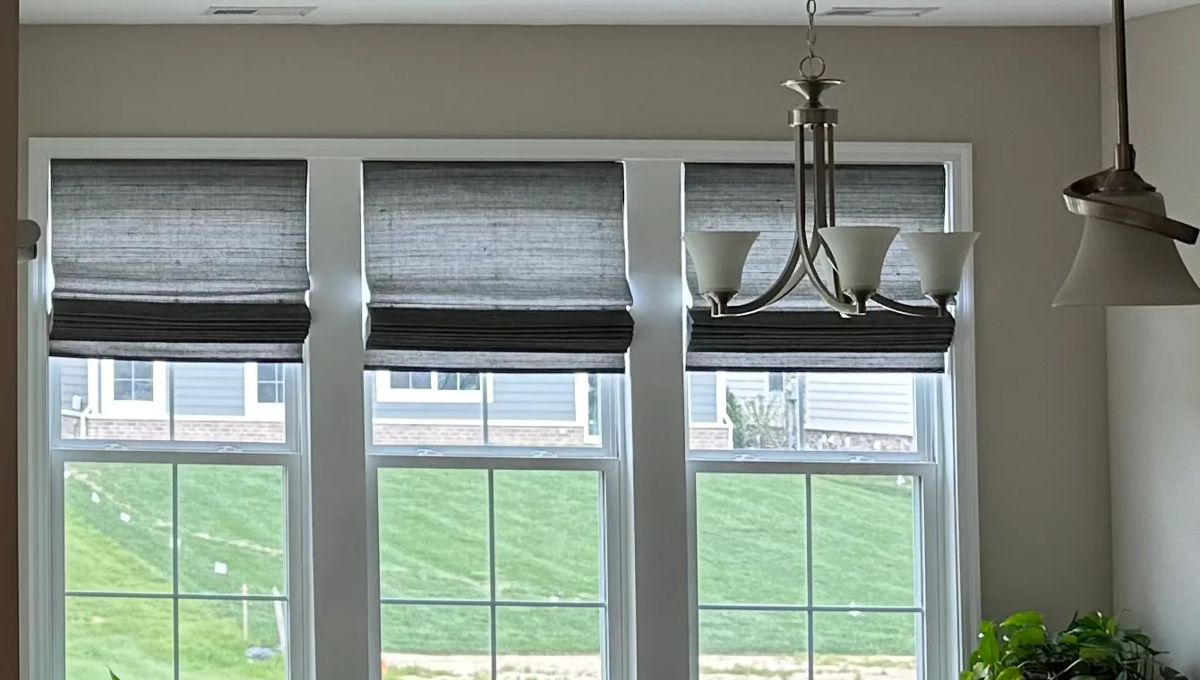 Three grey woven roman shades raised partway on a row of dining room windows, with a table and chairs below and a green lawn outside.