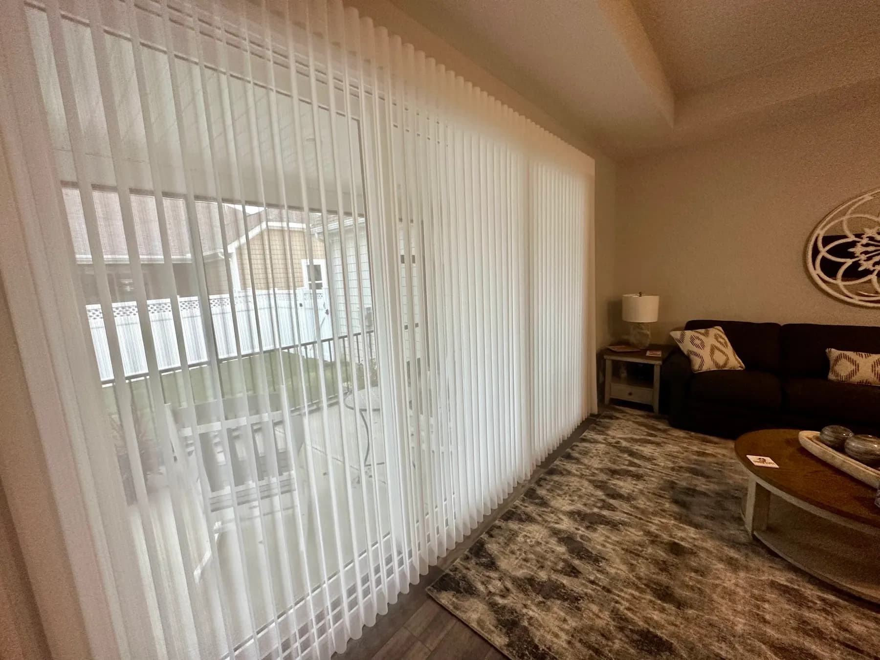 Floor-to-ceiling sheer vertical blinds drawn across a patio slider in a living room, with the backyard visible through the fabric vanes.