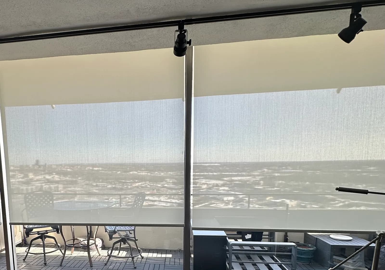 Two white solar screen shades lowered over balcony doors in a high-rise, with the city still visible through the weave.