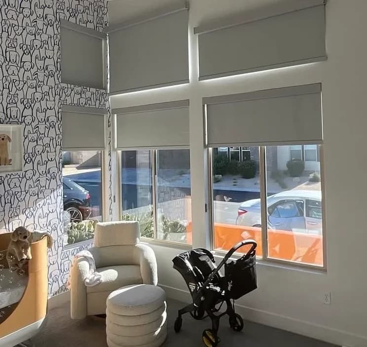 Light grey solar roller shades in white cassettes on tall corner windows of a nursery, lowered partway over a sunlit street, beside a rocking chair.