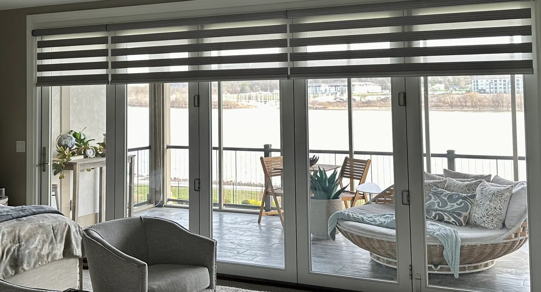 Charcoal transitional sheer shades raised across a four-panel glass slider wall in a living room, with a river and balcony seating visible beyond.