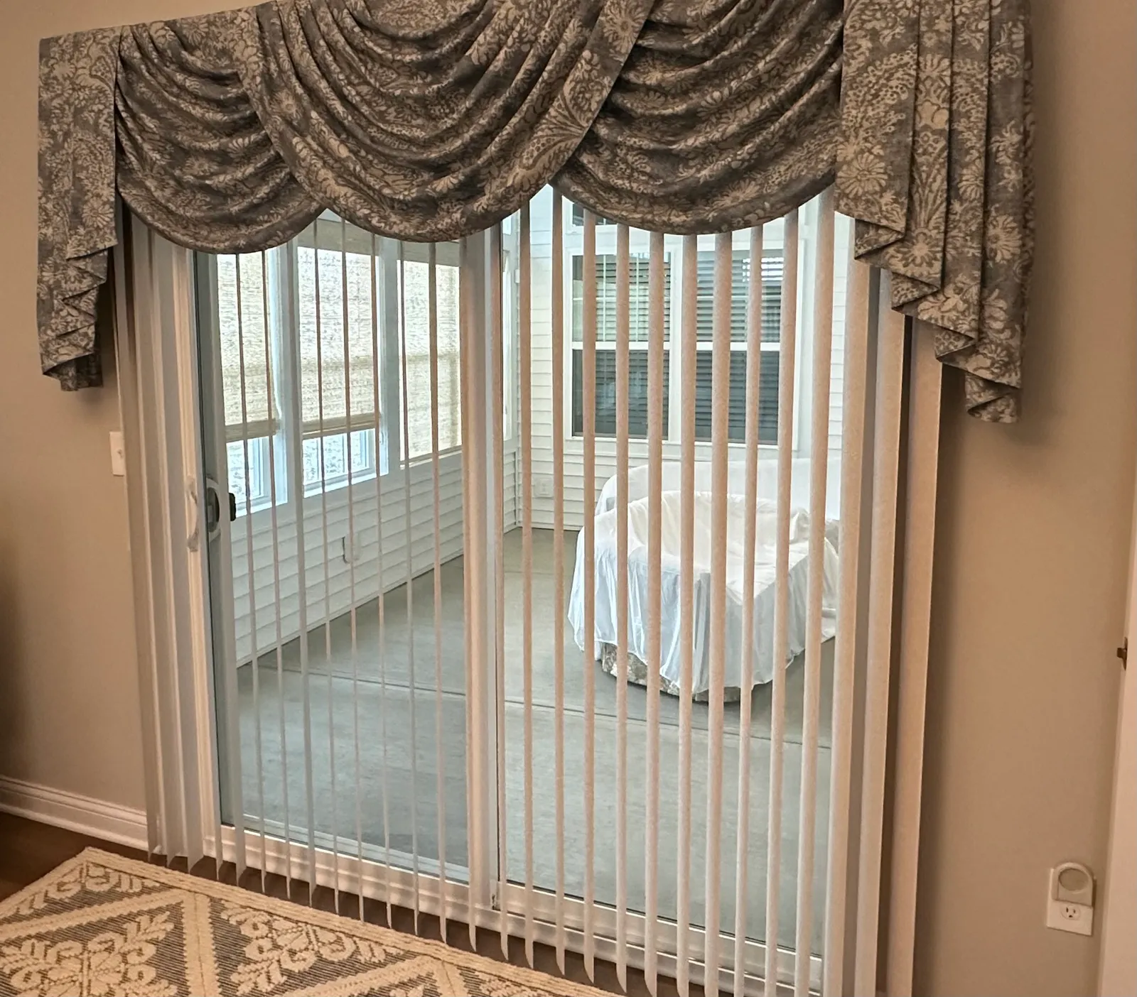 White vertical blinds drawn across a sliding patio door beneath a grey floral swag valance, with a sunroom visible through the glass.
