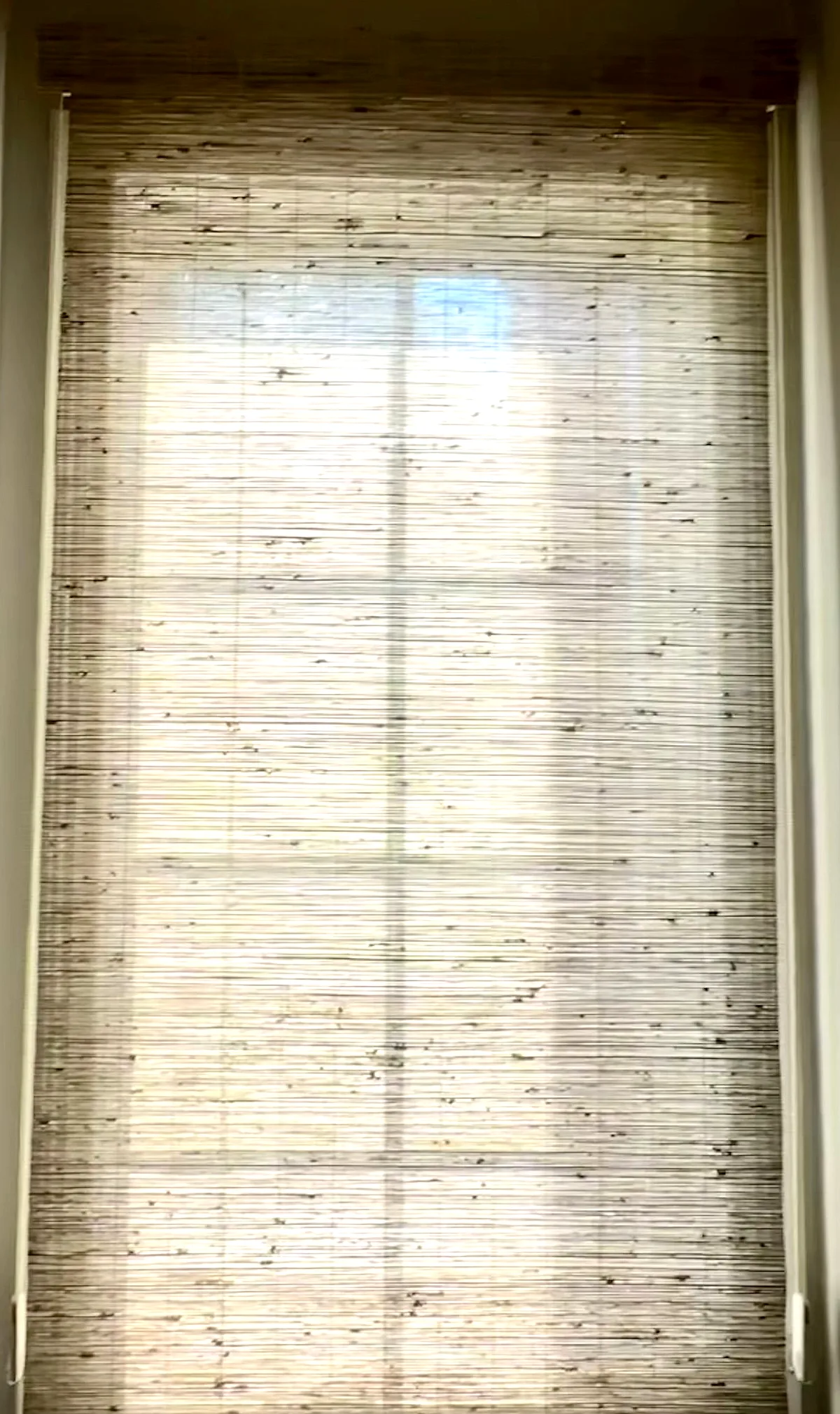 A natural woven wood shade lowered over a tall window at the end of a hallway, daylight glowing through the grass weave.