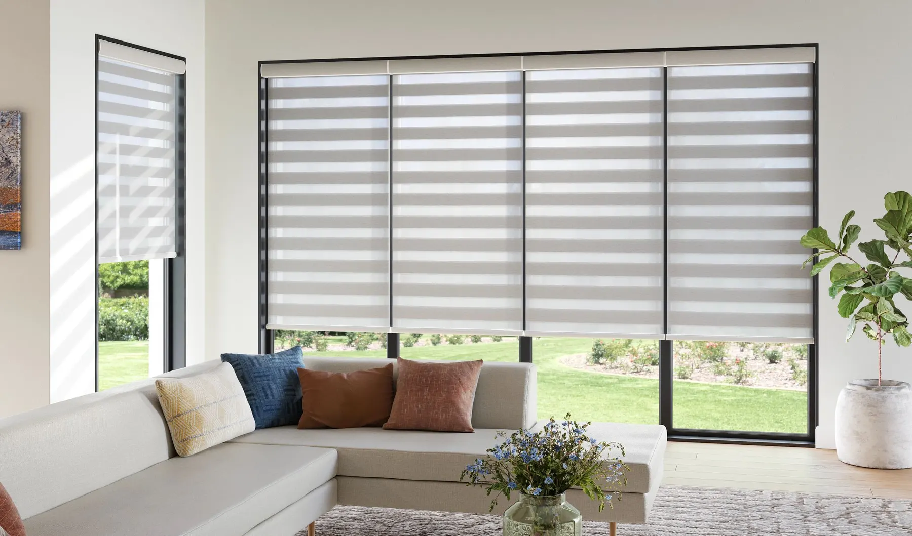Grey banded shades with alternating sheer and solid stripes across a wide picture window behind a white sectional sofa.
