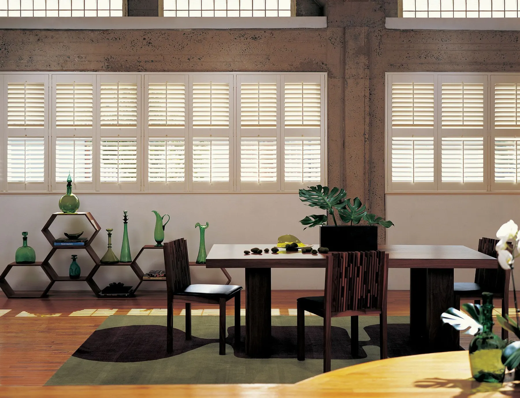 Cream plantation shutters across a long row of loft windows above a dark wood dining table.