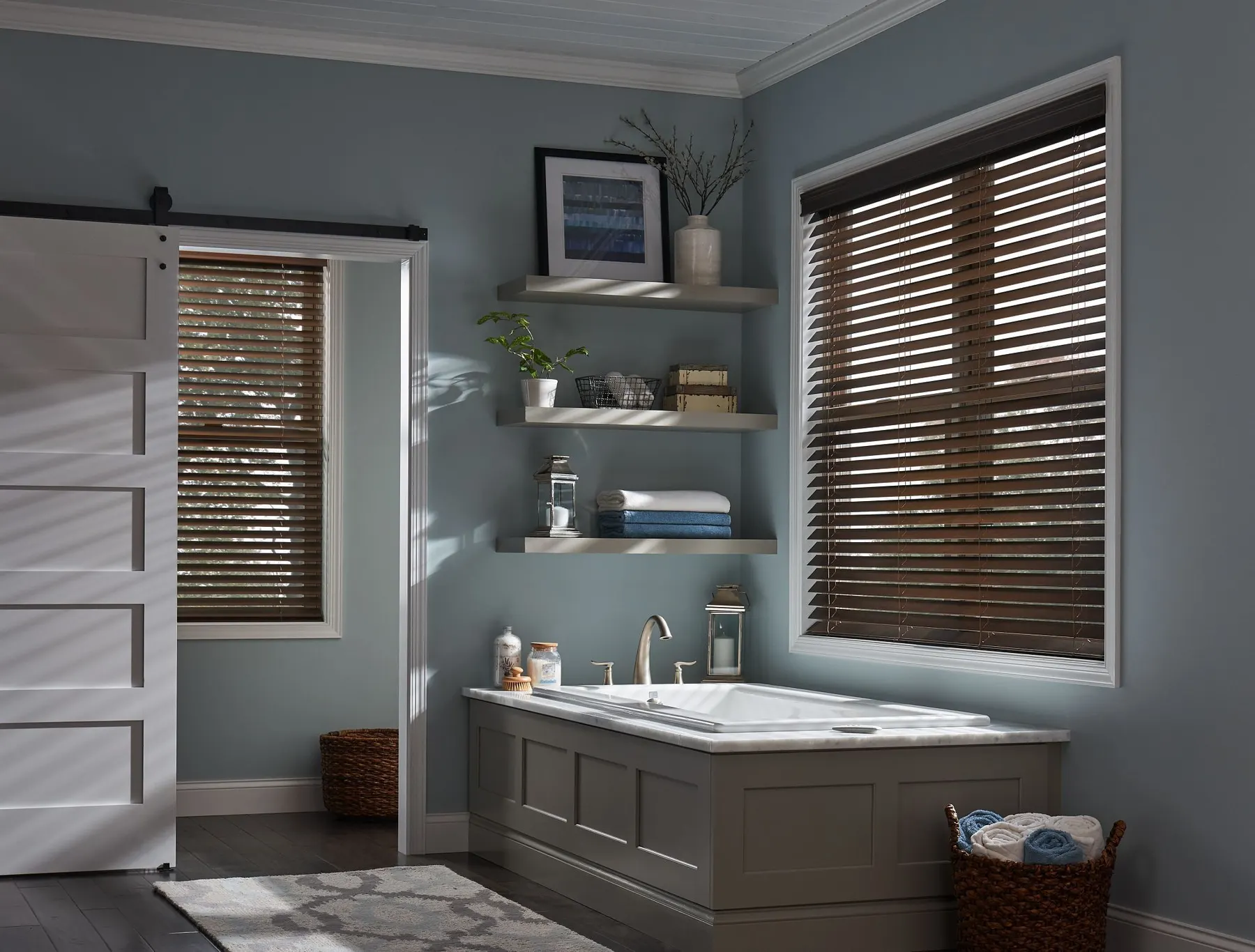 Dark faux wood blinds on two windows of a blue-grey bathroom with a soaking tub and a sliding barn door.