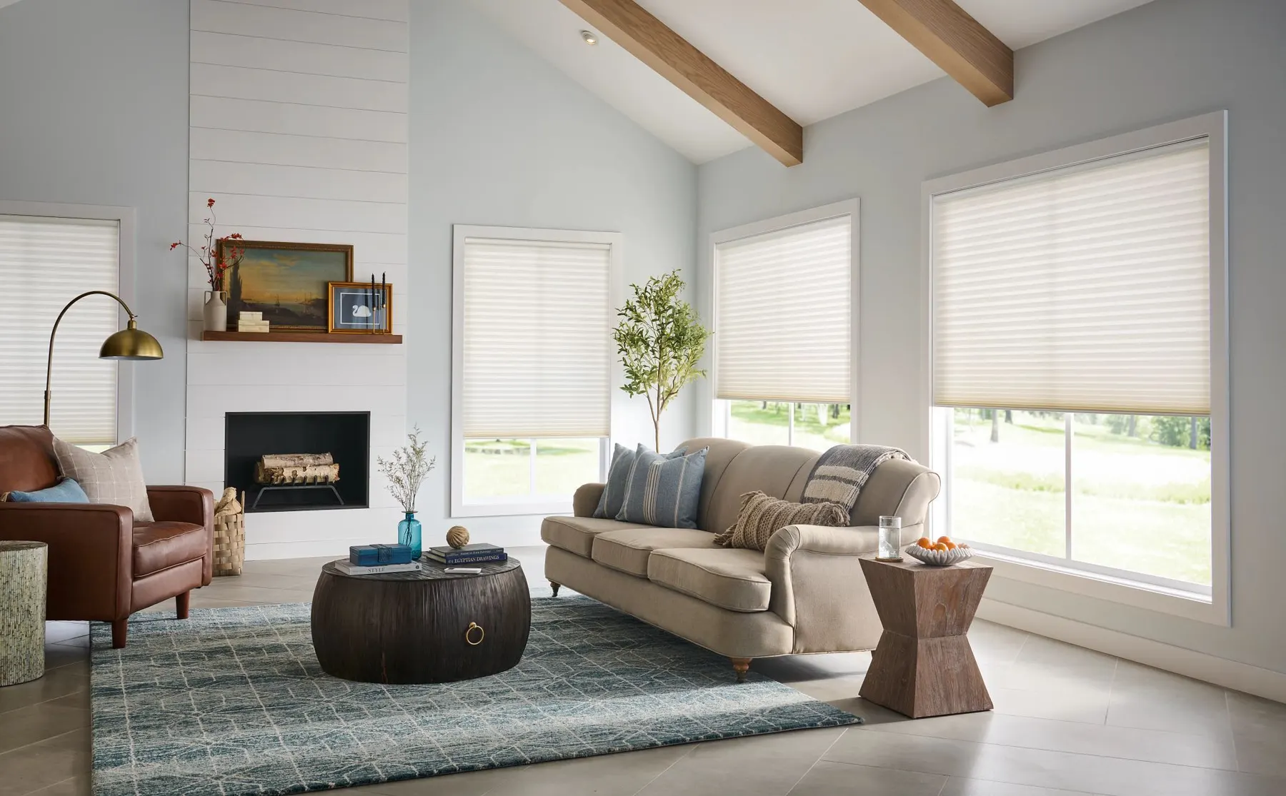 White honeycomb shades lowered partway on the windows of a bright living room with a vaulted, beamed ceiling and a fireplace.