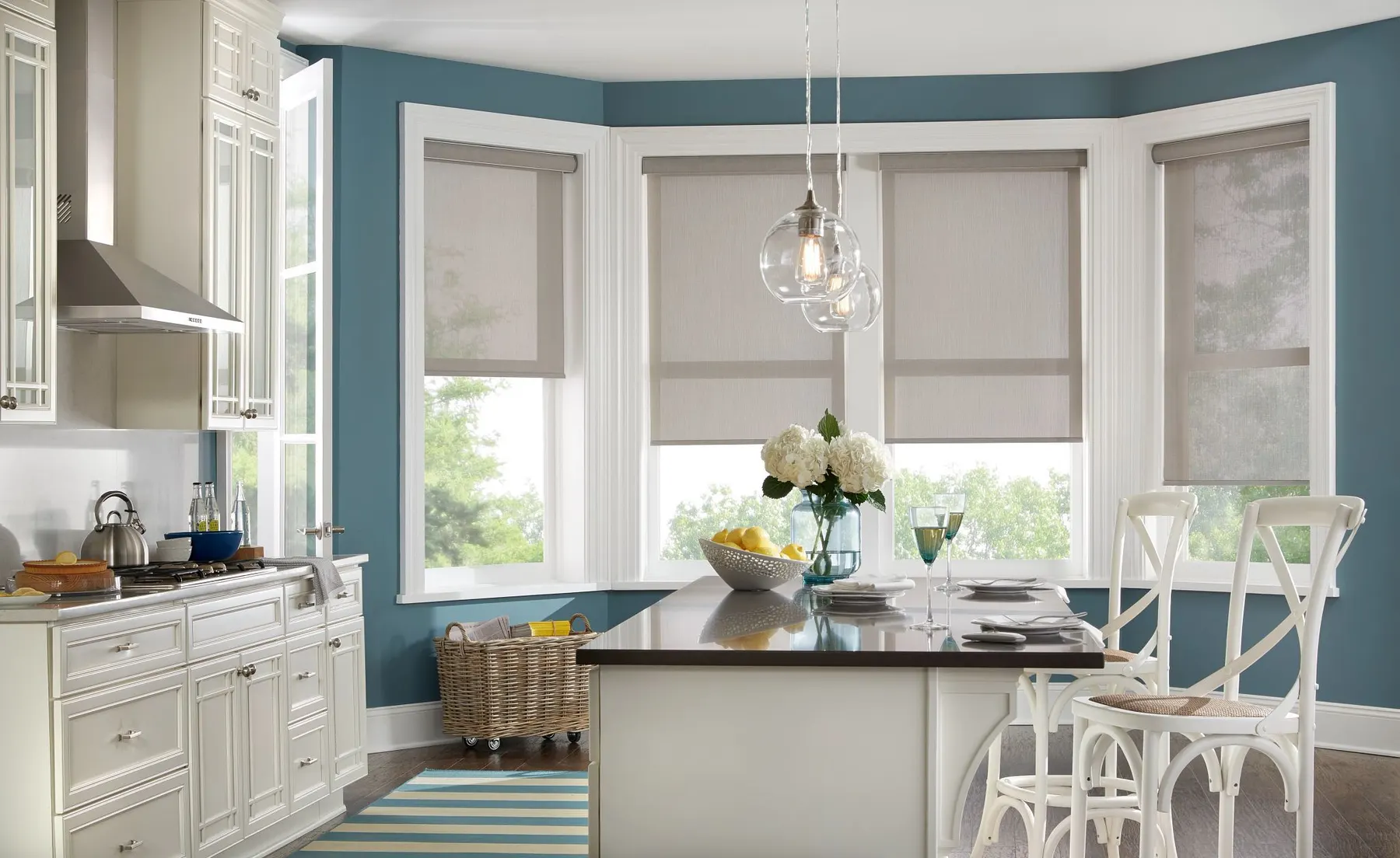 Taupe roller shades lowered halfway on a bay of kitchen windows behind a white island with bar stools.