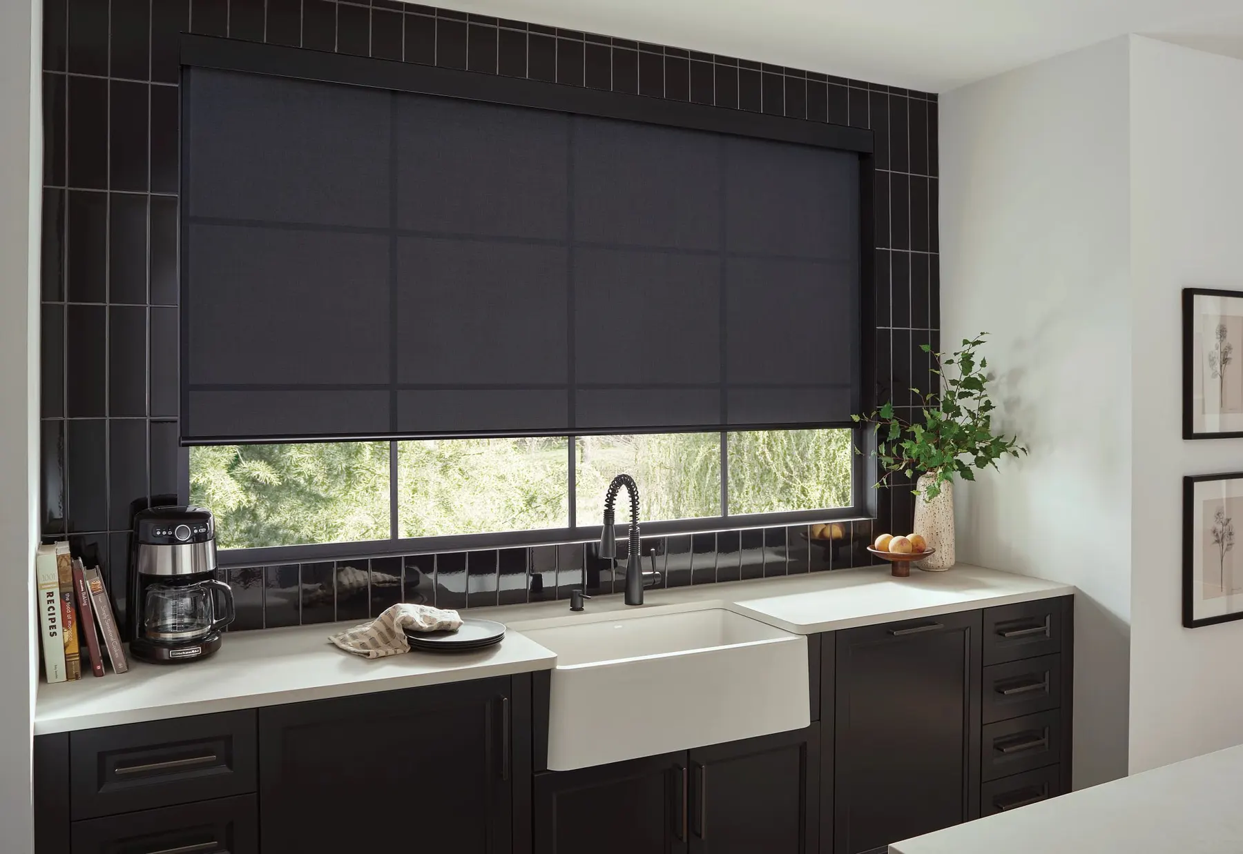 A wide charcoal roller shade lowered over a kitchen window above a white farmhouse sink and black tile wall.