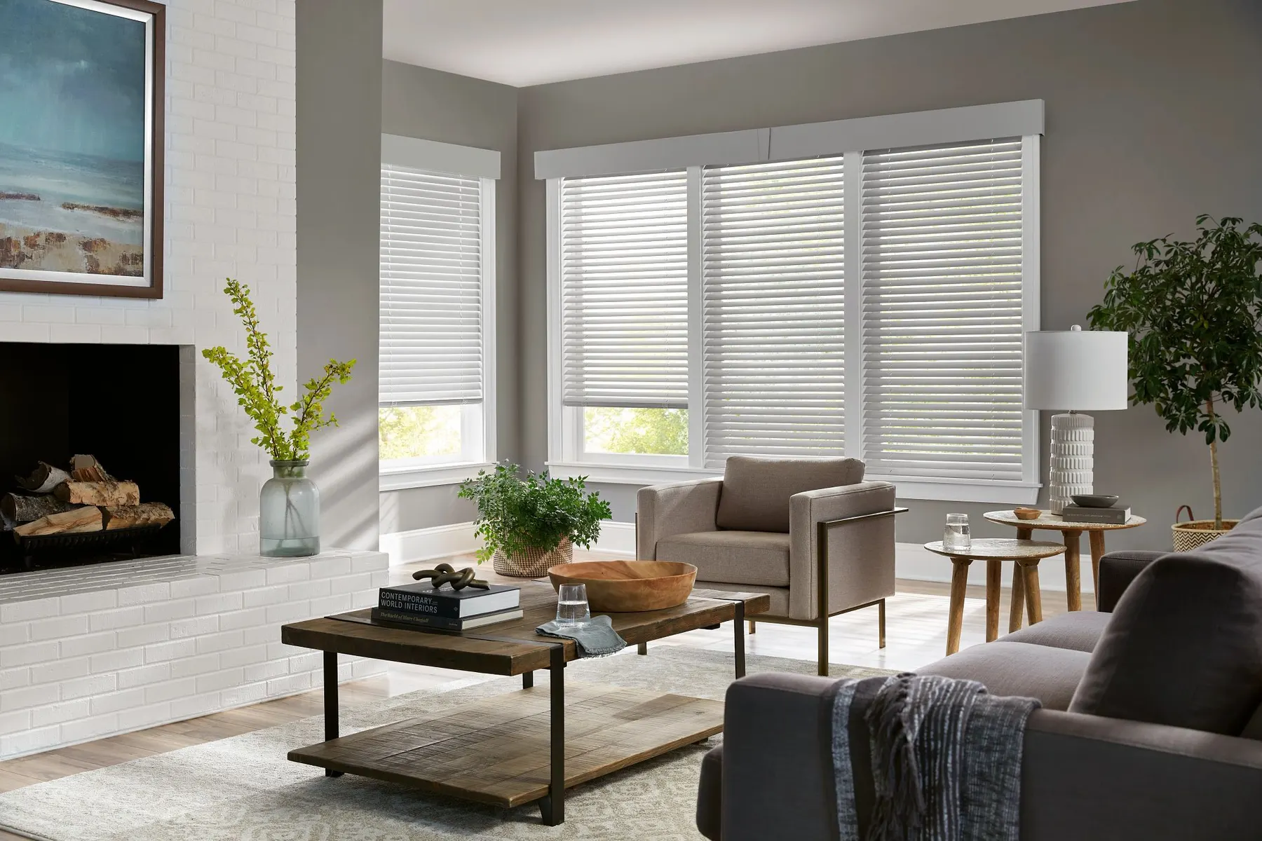 White wood blinds under matching cornices on a corner of windows in a living room with a white brick fireplace.