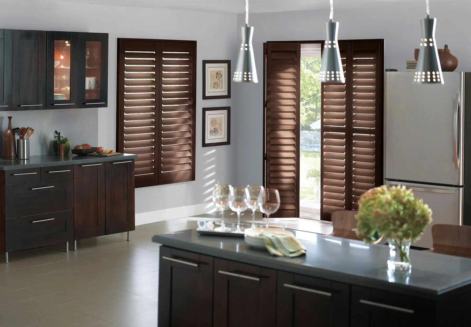 Dark stained wood shutters on a kitchen window and a pair of glass doors, beside espresso cabinets and pendant lights.