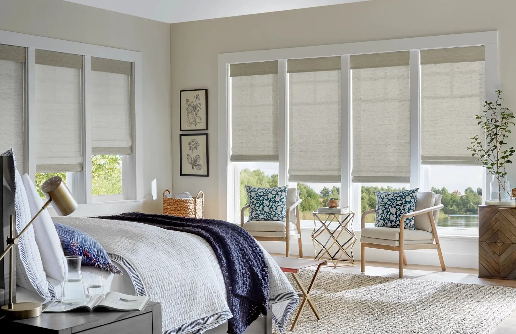 Natural woven shades on a wall of windows in a bright bedroom, with two armchairs looking out at the water.